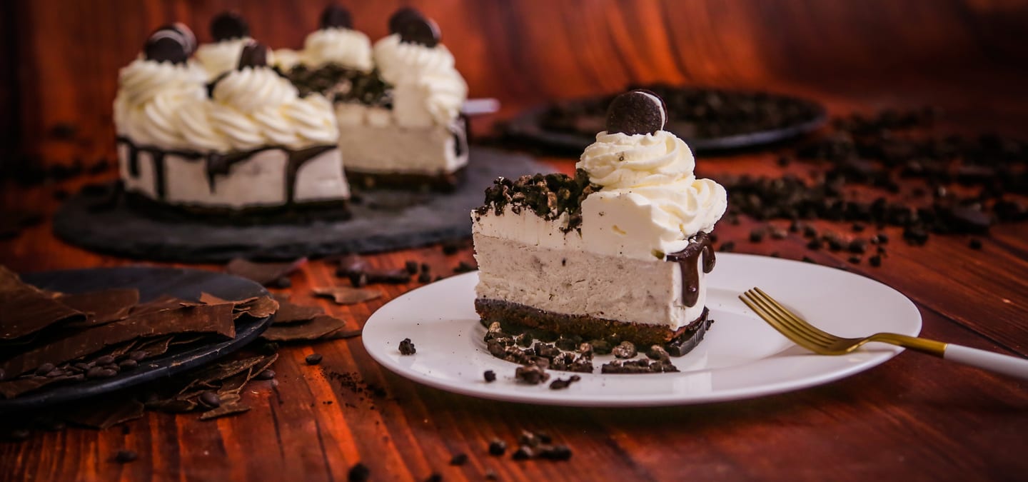 Tarta de queso Oreo: la pieza entera al fondo y una porción servida en plato, sobre una mesa de madera oscura